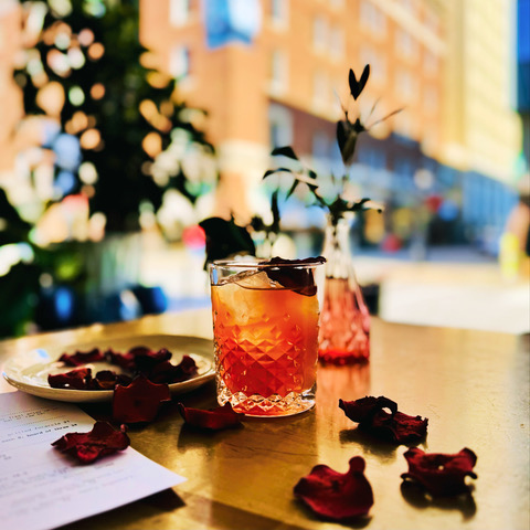Romantic cocktail garnished with a rose petal on a gold table, surrounded by scattered petals and elegant glassware, with a city view in the background.